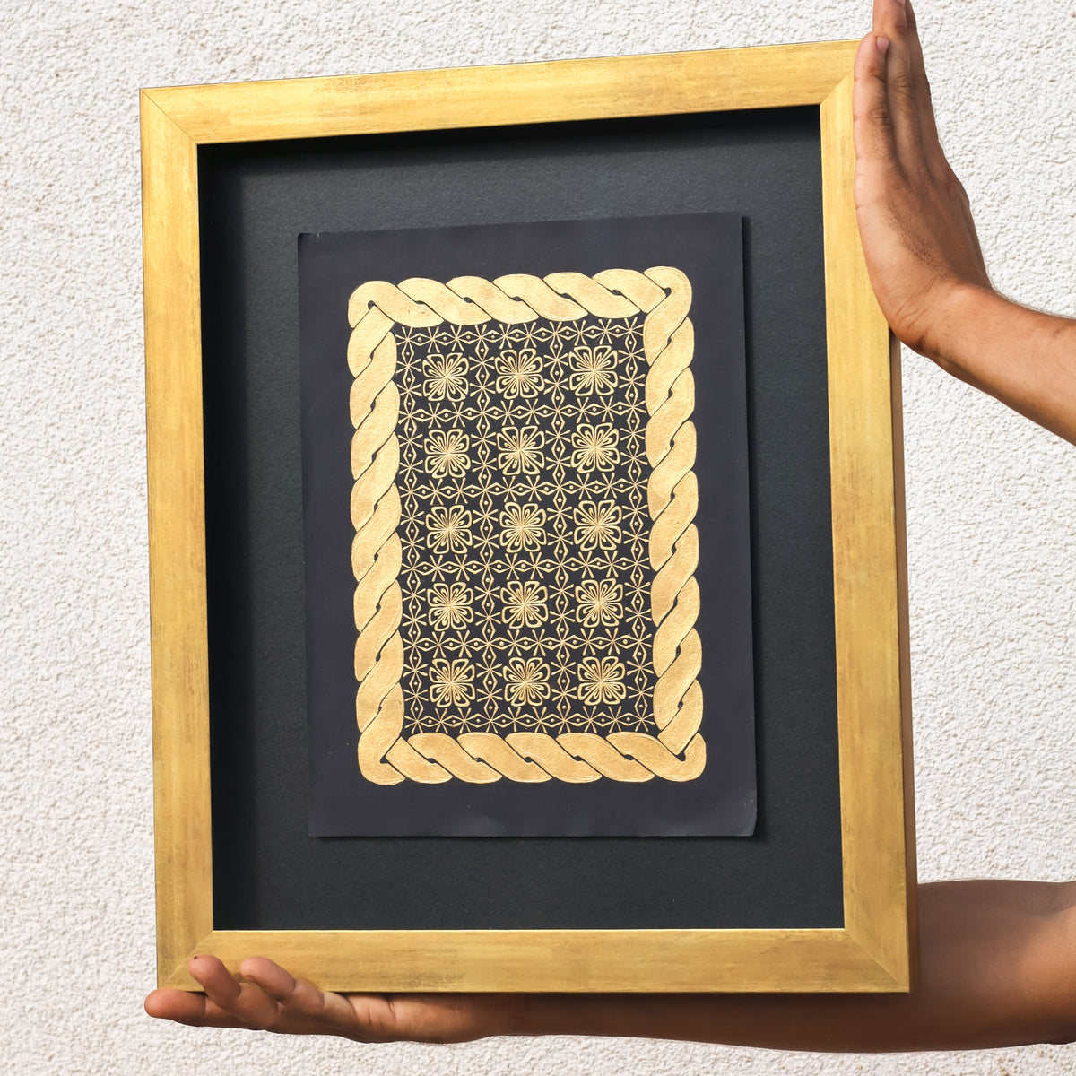 Framed artwork with gold geometric design on a black background, held by hands against a light gray wall.