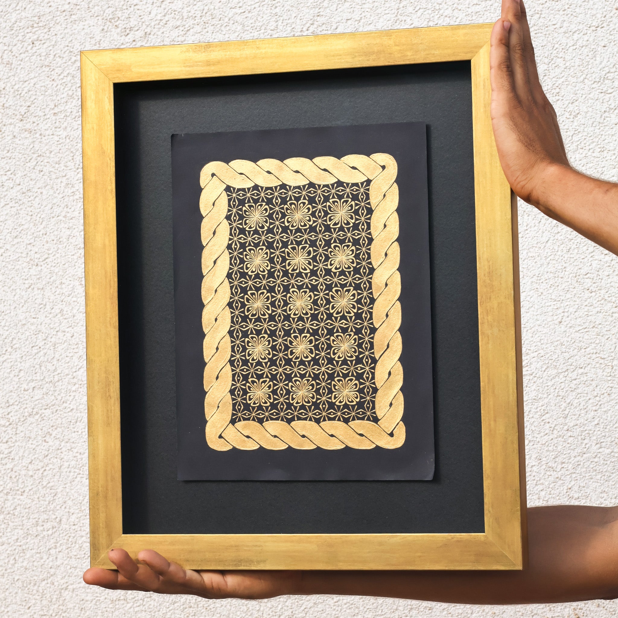 Framed artwork with gold geometric design on a black background, held by hands against a light gray wall.