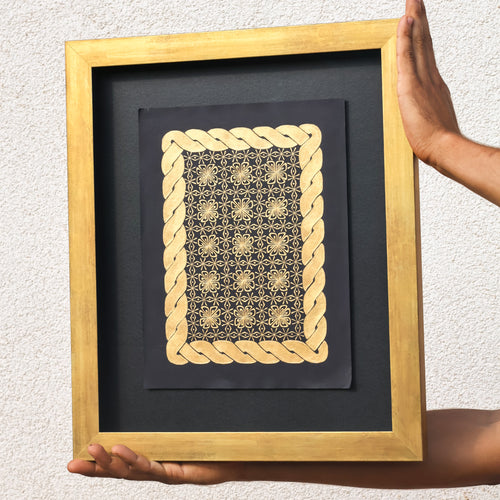 Framed artwork with gold geometric design on a black background, held by hands against a light gray wall.