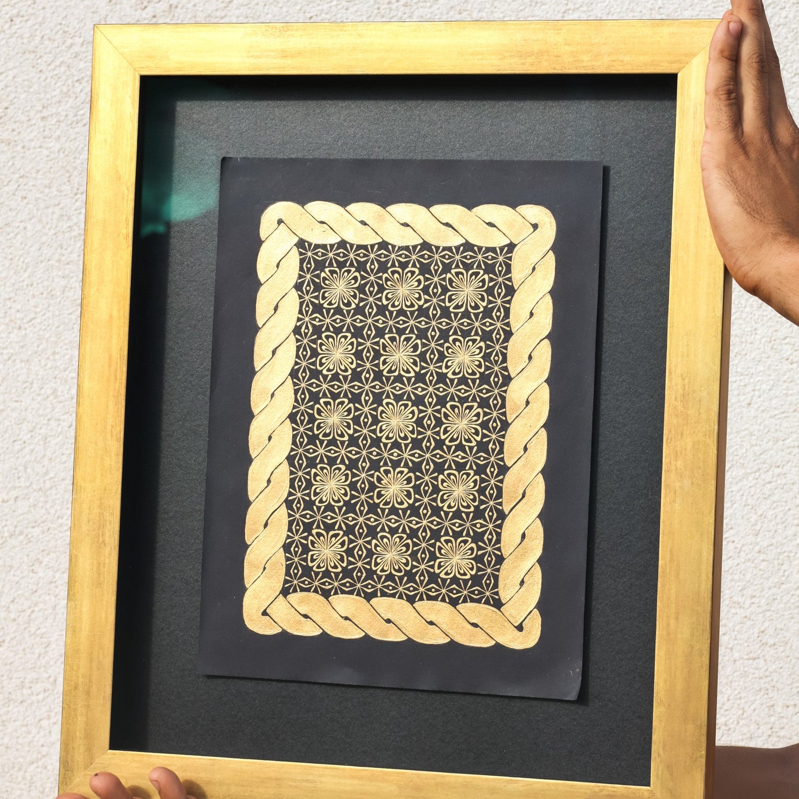 Framed artwork with gold geometric design on a black background, held by a person.