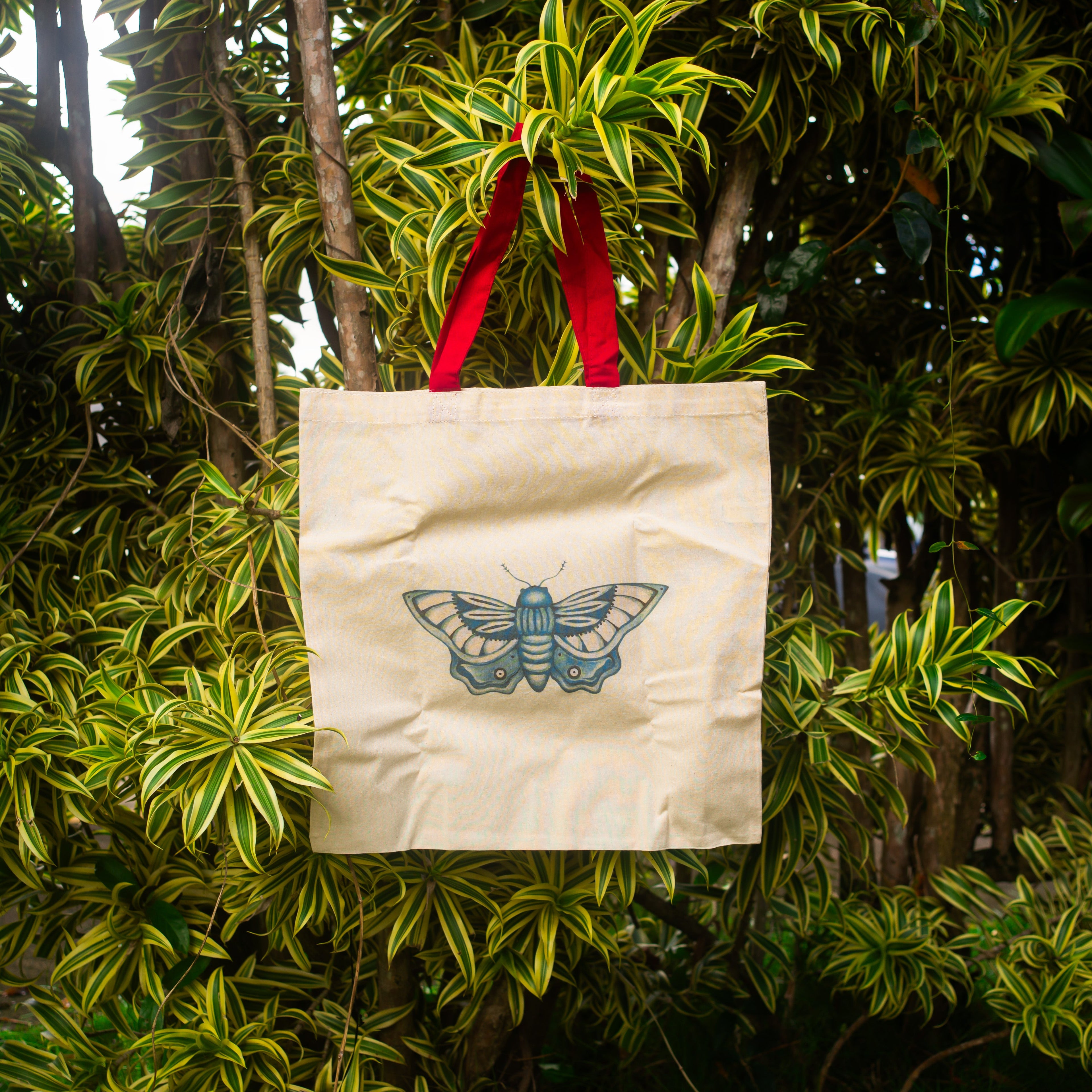 Cream canvas tote bag with red handles featuring a blue butterfly illustration, hanging from a leafy tropical tree with green and yellow foliage in natural outdoor light.